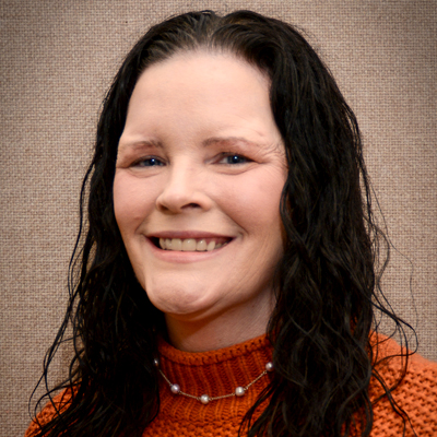 A person with long, wavy dark hair is wearing an orange knit sweater and a pearl necklace. The background is a textured beige surface.