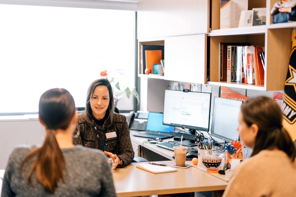 The image depicts a meeting or discussion taking place in an office setting. Three people are seated around a desk. Two of them are facing away from the camera, while one person, who appears to be leading the discussion, is facing towards the camera. The desk is cluttered with various items, including a coffee cup, pens, and papers. In the background, there are shelves filled with books and other office supplies, as well as two computer monitors displaying documents or spreadsheets.