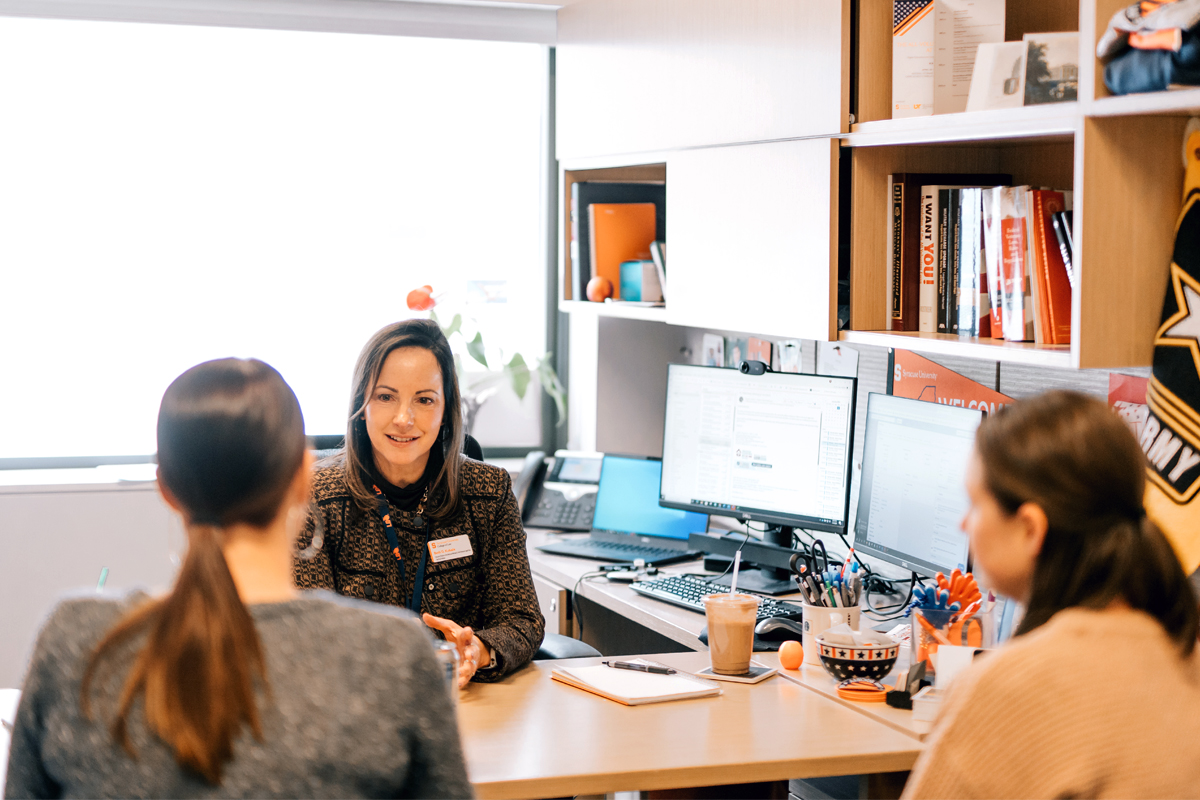 The image depicts a meeting or discussion taking place in an office setting. Three people are seated around a desk. Two of them are facing away from the camera, while one person, who appears to be leading the discussion, is facing towards the camera. The desk is cluttered with various items, including a coffee cup, pens, and papers. In the background, there are shelves filled with books and other office supplies, as well as two computer monitors displaying documents or spreadsheets.
