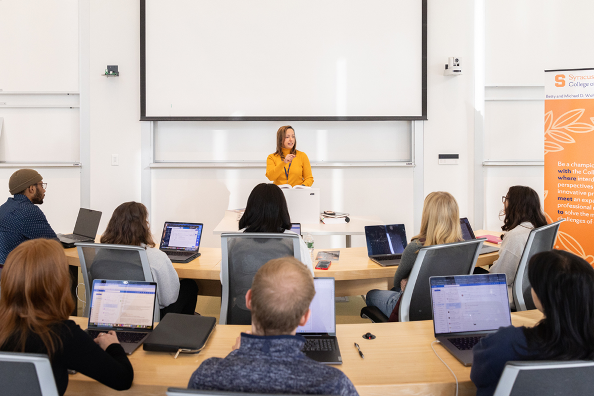 A classroom setting with a lecturer standing at the front, speaking to students who are seated and using laptops. The lecturer is positioned in front of a large projection screen. On the right side of the image, there is an orange banner that reads: "Syracuse College. Be a champion with our innovative programs that will help you succeed."