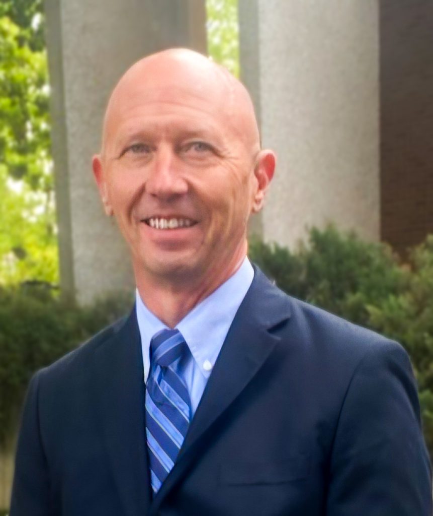 A person wearing a suit and tie standing outdoors, with their face blurred for privacy. The background features greenery and concrete structures.