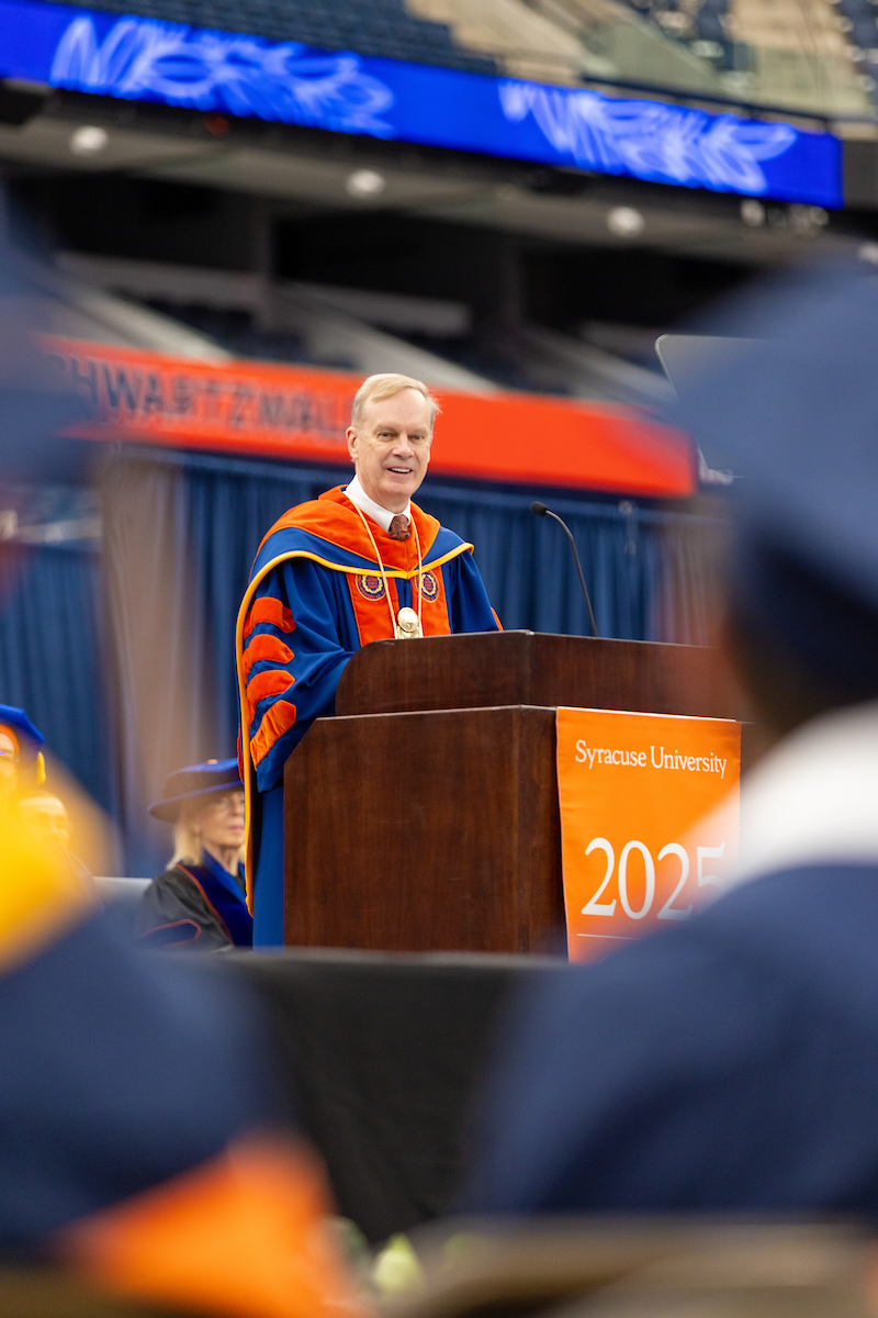 Chancellor Syverud, dressed in academic regalia, stands at a podium giving a speech. The podium has the text 'Syracuse University' and '2025' on it. The background includes other people in academic regalia.