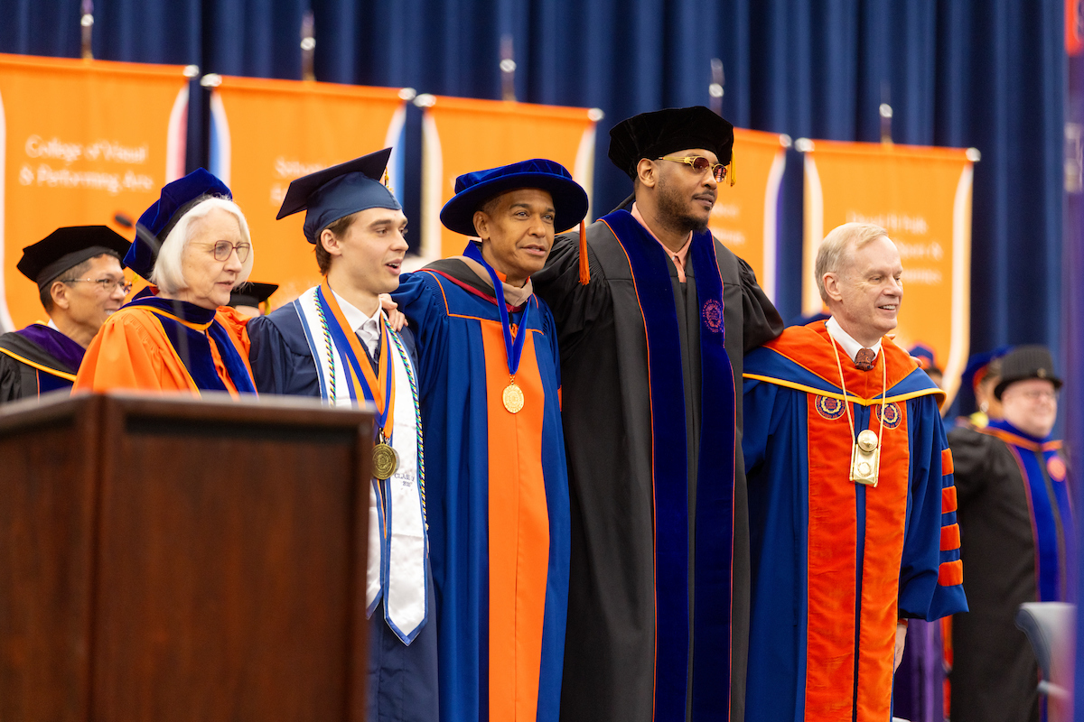A group of people dressed in academic regalia, standing in a row on a stage at Commencement 2025. There are banners in the background that include the names of schools/colleges.