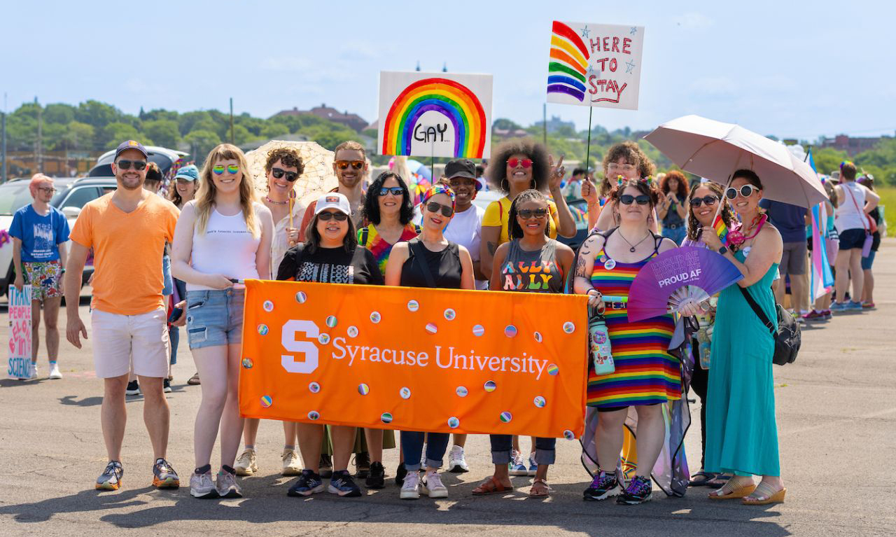 Group of people at the Central New York Pride Parade, holding an orange 'Syracuse University' banner, with rainbow-colored signs including one that says 'GAY' and another that says 'HERE TO STAY."