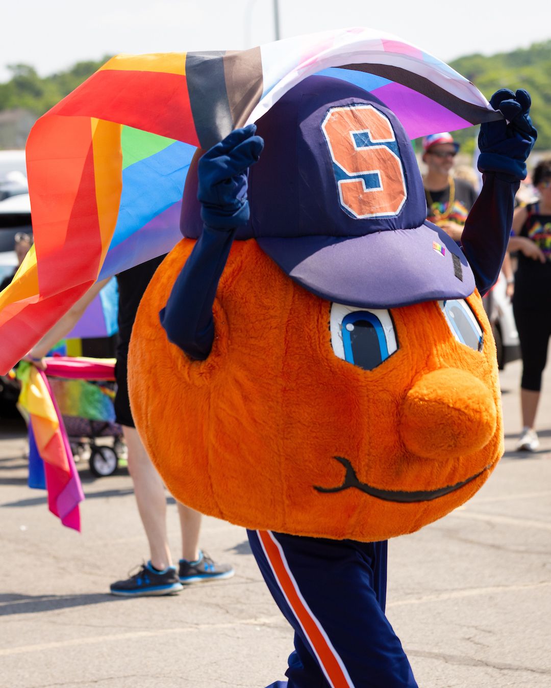 Syracuse University mascot Otto the Orange holds a colorful pride flag above their head, with people and other flags in the background, at a Pride month event.