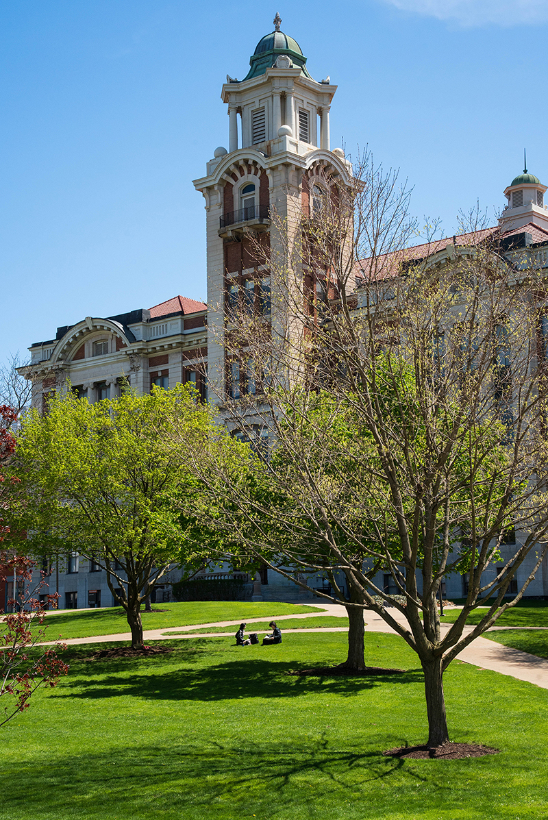A large historic building featuring a tall clock tower and dome, surrounded by trees and a manicured lawn with pathways. Several people are sitting on the grass in the foreground.