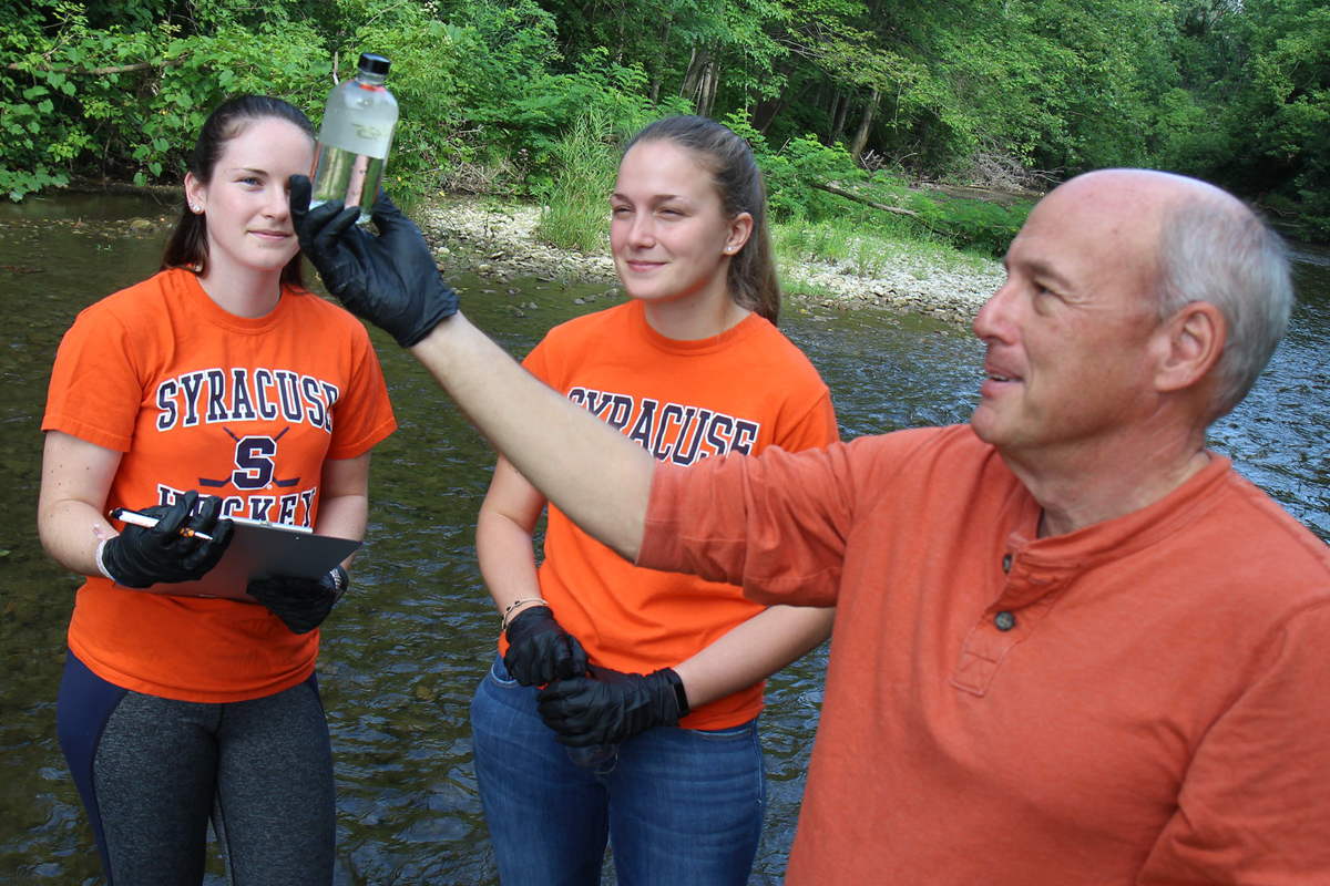 Three people are standing in a shallow river surrounded by greenery. Two of them are wearing orange "Syracuse" t-shirts, and one is holding a clipboard. The third person, wearing an orange long-sleeve shirt, is holding up a small clear bottle filled with water. 