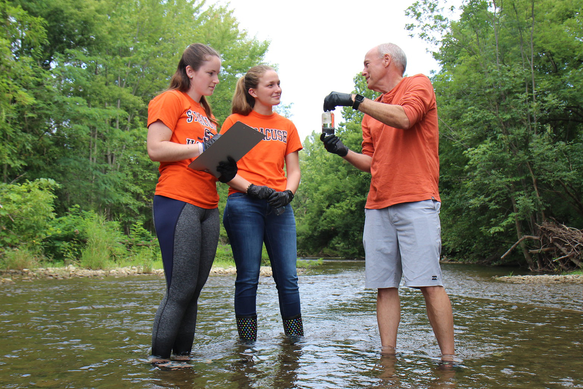 The image shows three people standing in a shallow stream surrounded by lush green trees. Two of the individuals are wearing orange shirts and holding a clipboard, while the third person is also wearing an orange shirt and appears to be explaining something, gesturing with gloved hands. The scene suggests an outdoor educational or research activity.