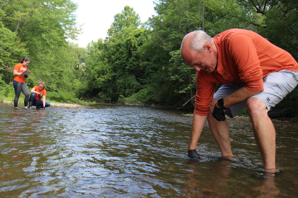 The image shows three people in a shallow stream surrounded by lush green trees. The person in the foreground, wearing an orange long-sleeve shirt and gray shorts, is bending down and touching the water with gloved hands. Two other people are in the background, also wearing orange shirts; one is standing while the other is crouching near the water. The scene appears to be set in a natural environment with clear water and dense foliage on both sides of the stream.