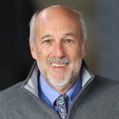 The image shows a person wearing a grey zip-up jacket over a blue shirt and a colorful patterned tie. The background is dark and out of focus.
