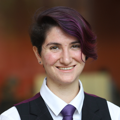 A person with short, dark hair that has purple highlights is wearing a white dress shirt, a purple tie, and a black vest. The background is blurred and appears to be indoors.