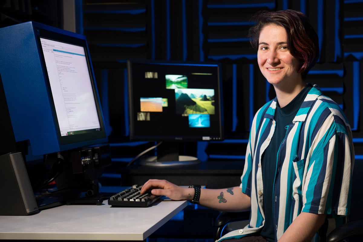 A person wearing a striped shirt with tattoos on their arm is sitting at a desk in front of two computer monitors. The monitor on the left displays text, while the monitor on the right shows several images of landscapes. The background features soundproofing panels.