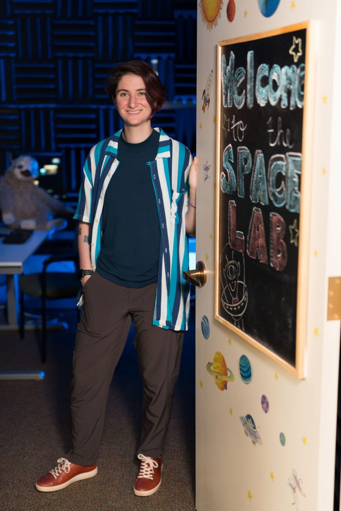 A person is standing in a doorway, leaning against the door frame. The door has a blackboard with colorful chalk writing that says 'Welcome to the SPACE LAB' along with drawings of planets and stars. The room behind the person has acoustic foam panels on the walls and a large stuffed animal sitting at a table.