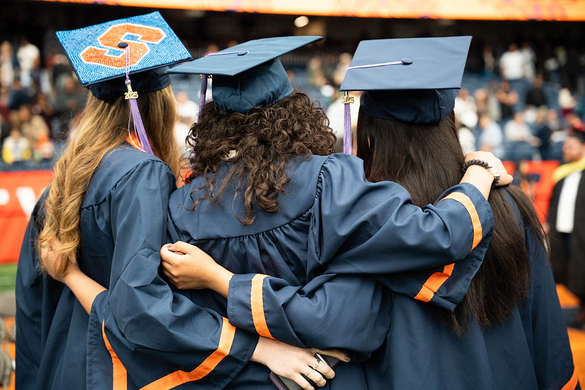 Three graduates in blue caps and gowns stand with their backs to the camera, arms around each other. The graduate on the left has a decorated cap with an orange 'S' on it. They are in a stadium, with blurred spectators in the background.
