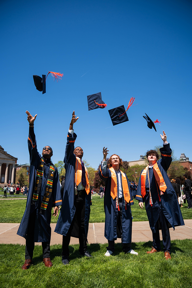 Four individuals wearing graduation gowns toss their caps in the air while standing on a grassy area of the Quad with a building in the background.