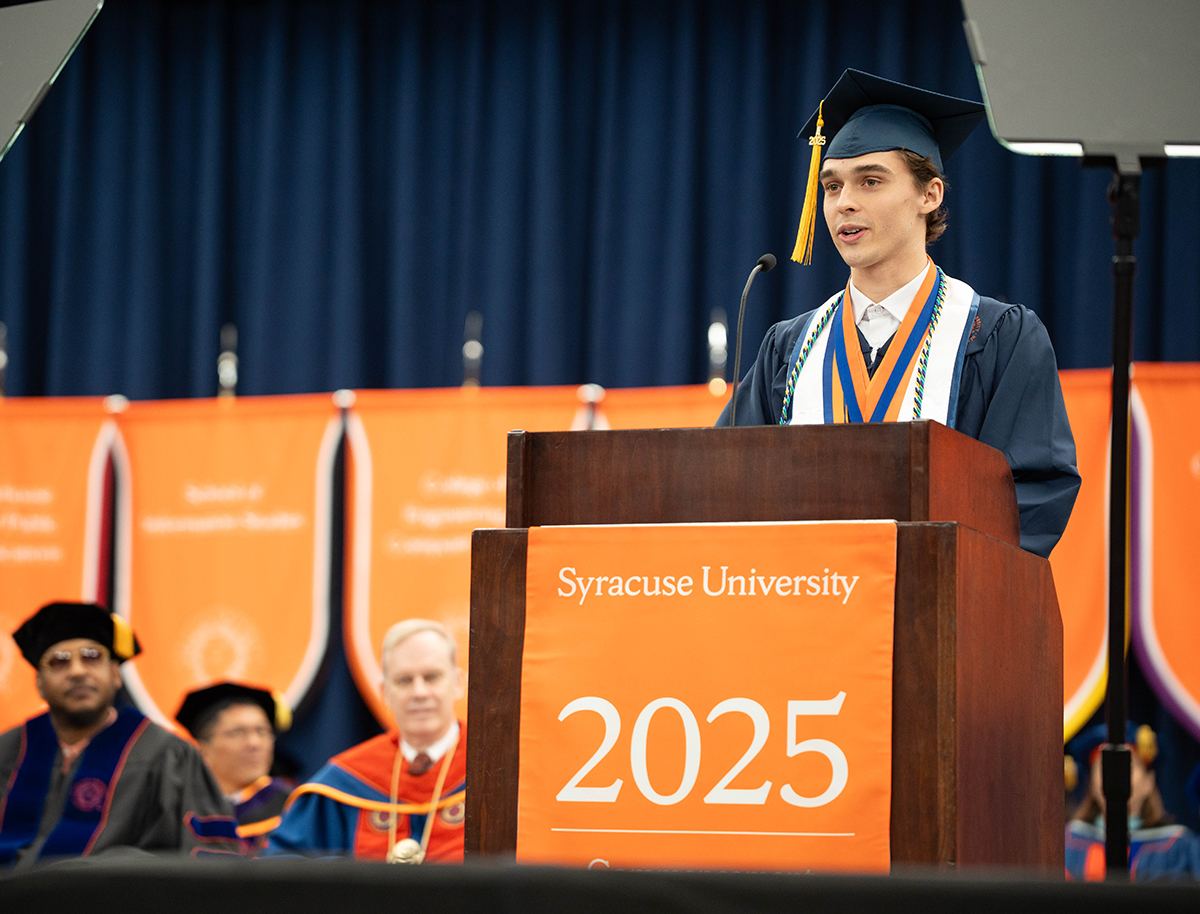 A graduate in a cap and gown stands at a podium with the text 'Syracuse University 2025' on it. Several people are seated behind the podium, also in academic regalia, with orange banners and white text hanging in the background.