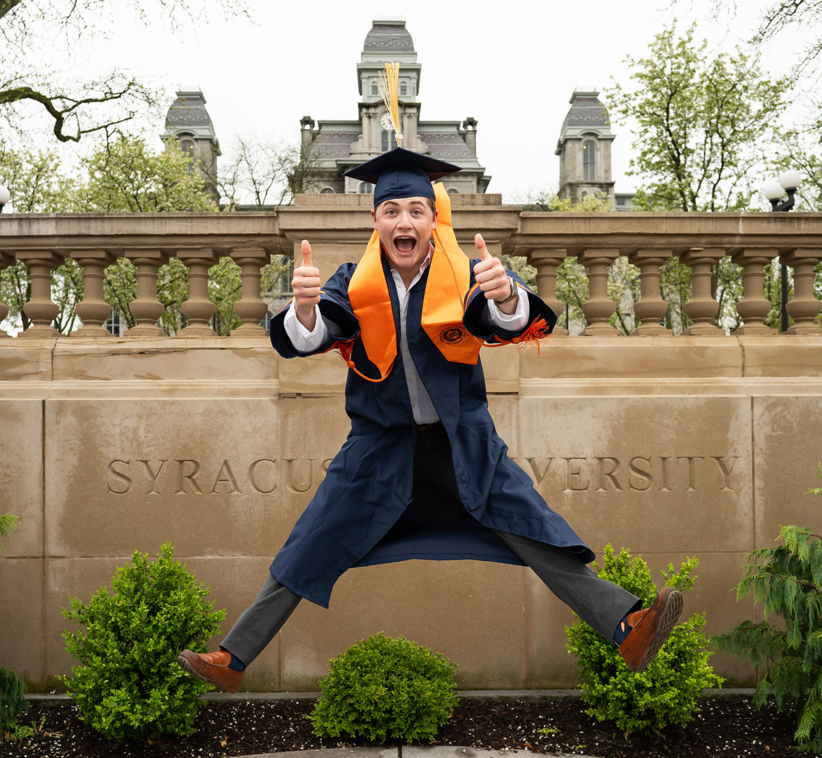 A person in graduation attire, including a cap and gown, jumping in the air with both thumbs up. The background features a stone wall with the engraved text 'SYRACUSE UNIVERSITY' and some greenery. The Hall of Languages can be viewed in the background at a distance.