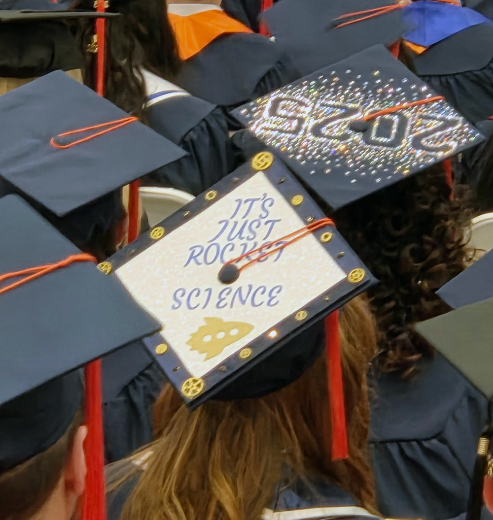 A group of graduates wearing caps and gowns, with the focus on their decorated graduation caps. One cap has the text 'IT'S JUST ROCKET SCIENCE' with a small rocket illustration, and another cap has the year '2025' written in glittery decorations.