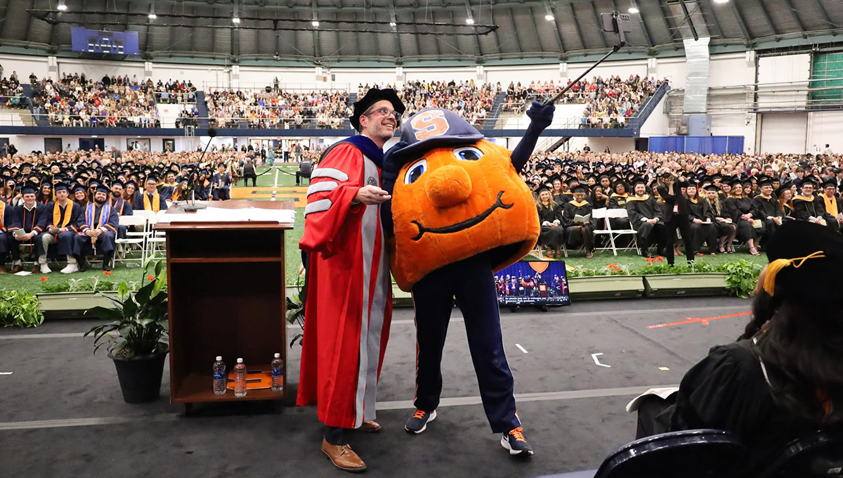 At the Falk College convocation ceremony, Dean Jeremy Jordan dressed in academic regalia stands next to Otto the Orange, taking a selfie with the crowd of graduates and attendees in the background. The stage has plants and water bottles on it, and the audience is seated in rows behind the stage.