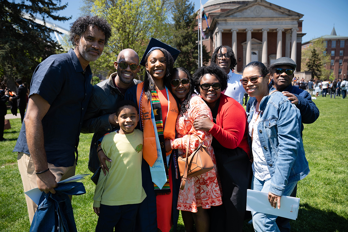 A group of people stand together on a grassy area in front of a large building with columns at Commencement 2025. The individuals are dressed in various outfits, with one person wearing a graduation cap and gown.