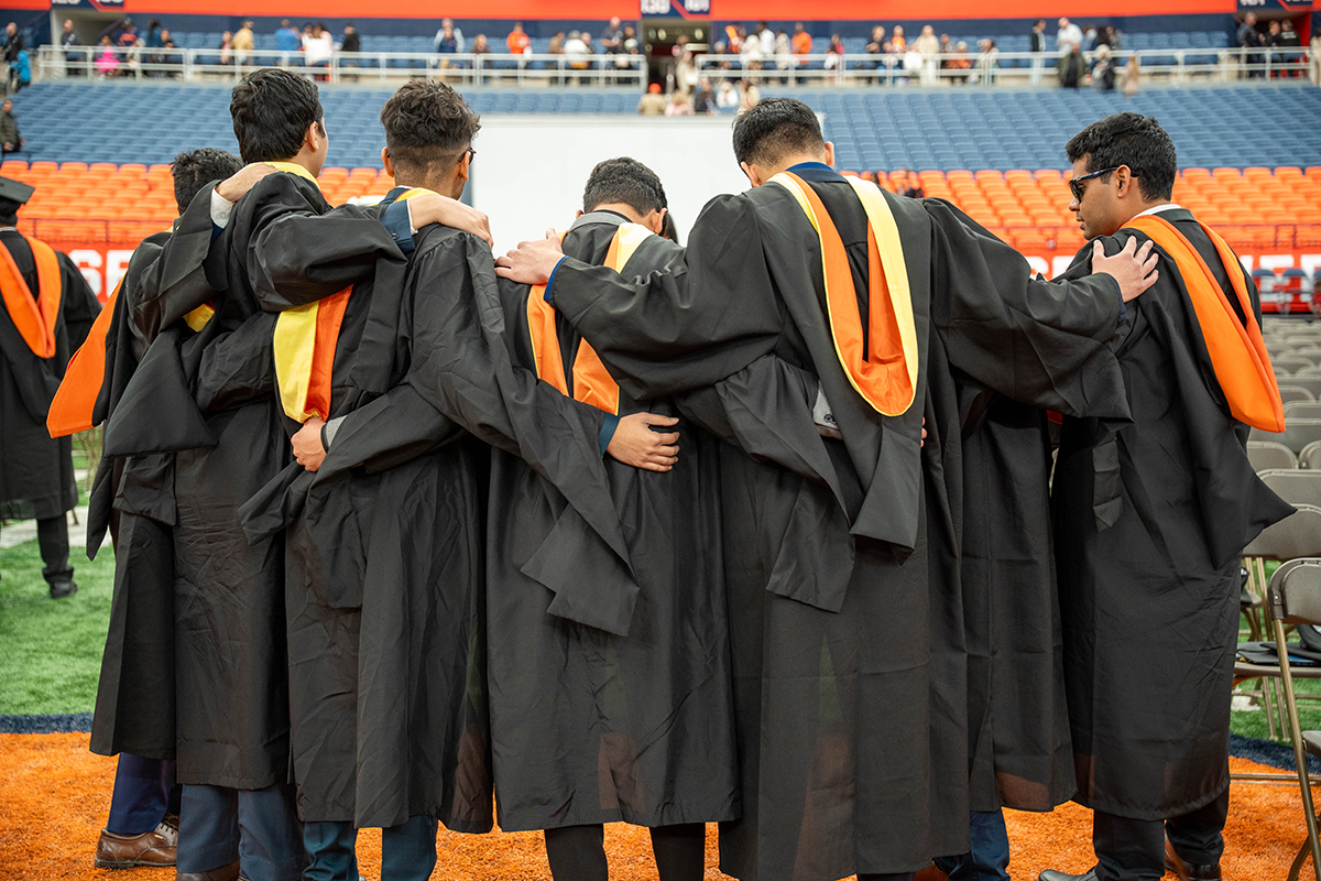 A group of people wearing graduation gowns and caps, stand in a row with their arms around each other. They are facing away from the camera, and the setting is a stadium with rows of seats in the background.
