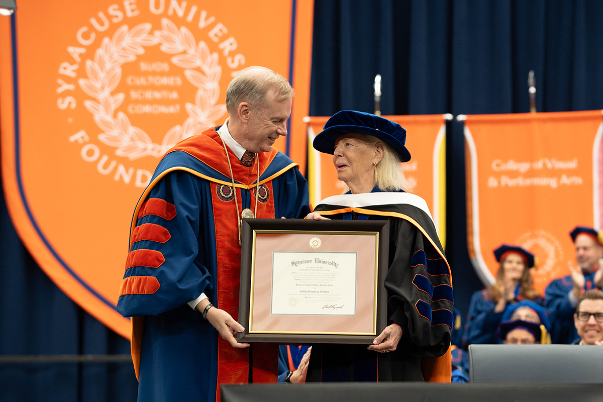 Two individuals in academic regalia, Chancellor Kent Syverud and Judith Greenberg Seinfeld, hold a framed diploma from Syracuse University.