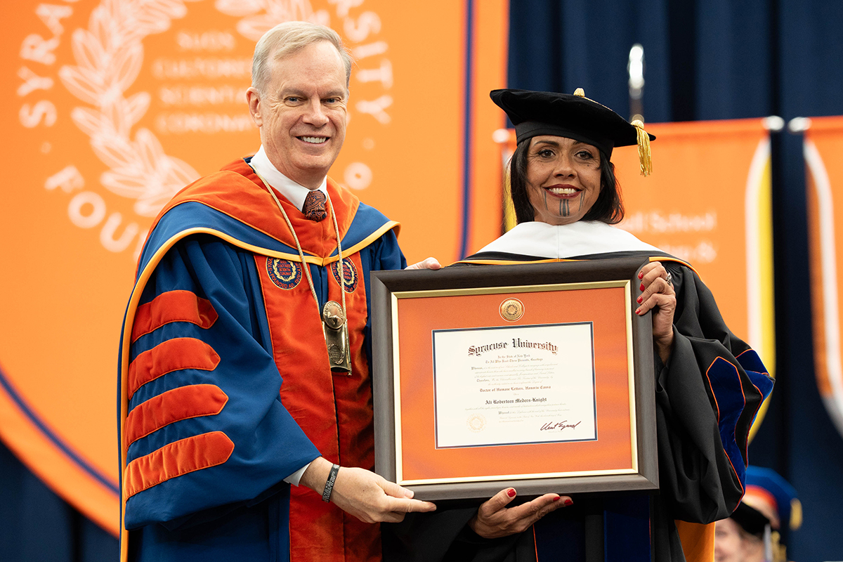 Two individuals in academic regalia, Chancellor Kent Syverud and Ali Meders-Knight, hold a framed diploma from Syracuse University.