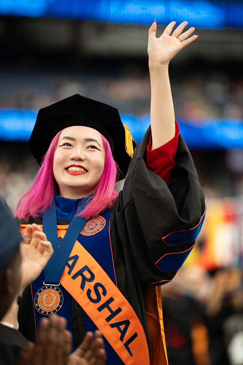 A person wearing graduation attire, including a cap and gown, with a sash that reads 'MARSHAL.' The person is raising one hand in the air and smiling.