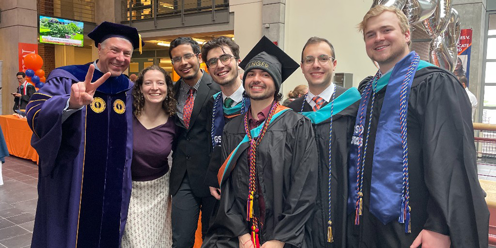 A group of 7 people dressed in graduation attire, standing together indoors. Some are wearing caps and gowns with various colored sashes and cords, indicating different academic achievements or honors. The person on the left, Maxwell Dean David Van Slyke, is making a peace sign with their hand. The background includes a table with orange and blue decorations and balloons, as well as other people and architectural elements of the building.