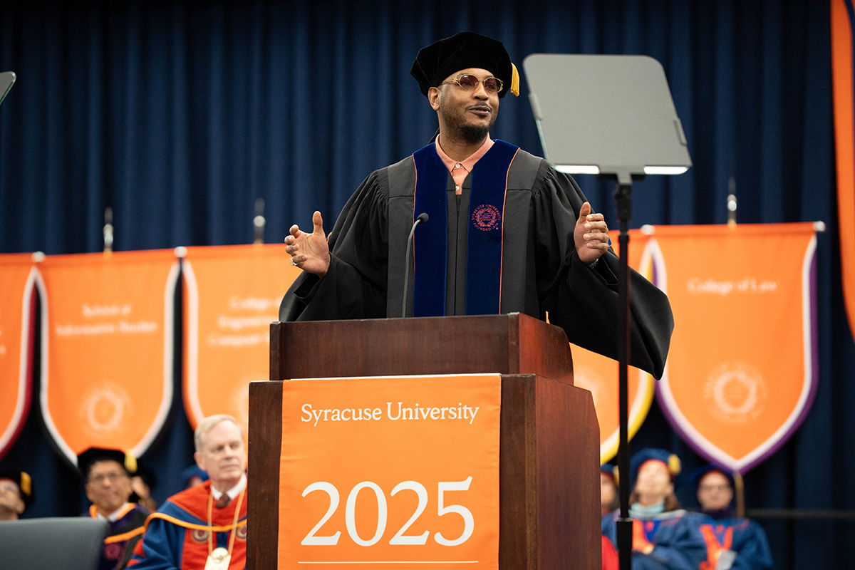 Carmelo Anthony in academic regalia speaks at a podium during the Syracuse University 2025 commencement ceremony. The podium has an orange banner with the text 'Syracuse University 2025.' In the background, there are several other individuals in academic regalia seated, and orange banners with various college names.