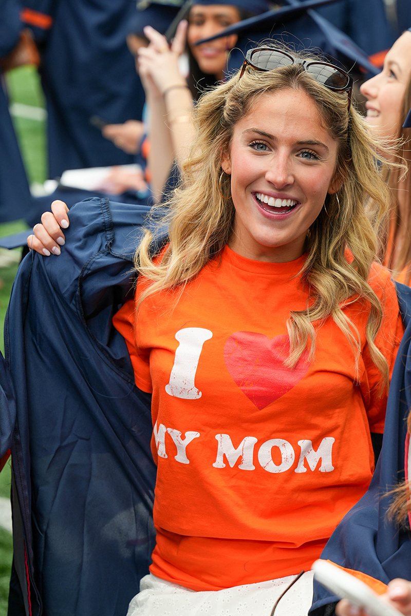 A person wearing an orange t-shirt with the text 'I LOVE MY MOM' printed on it. The person is holding open a dark blue graduation gown, revealing the t-shirt underneath. There are other people in the background also wearing graduation gowns.