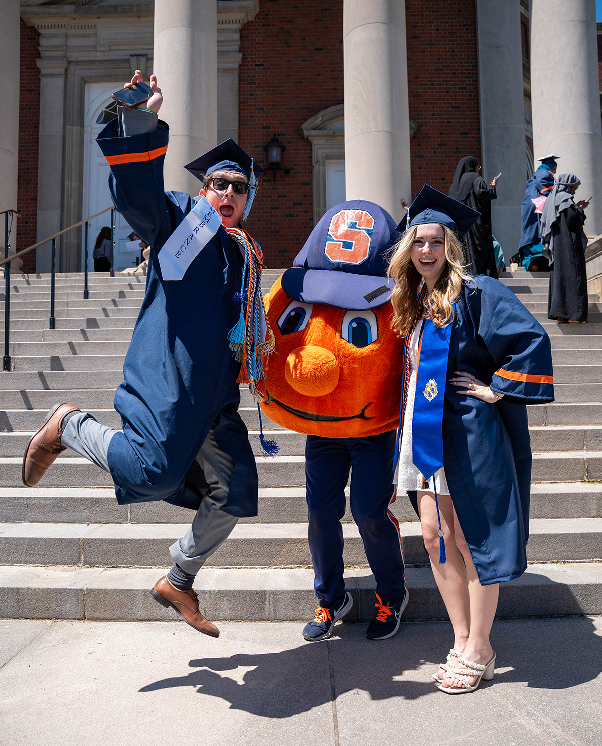 Two individuals in graduation attire stand on the steps of a building with large columns. One individual is jumping in the air and Otto the Orange is between them.