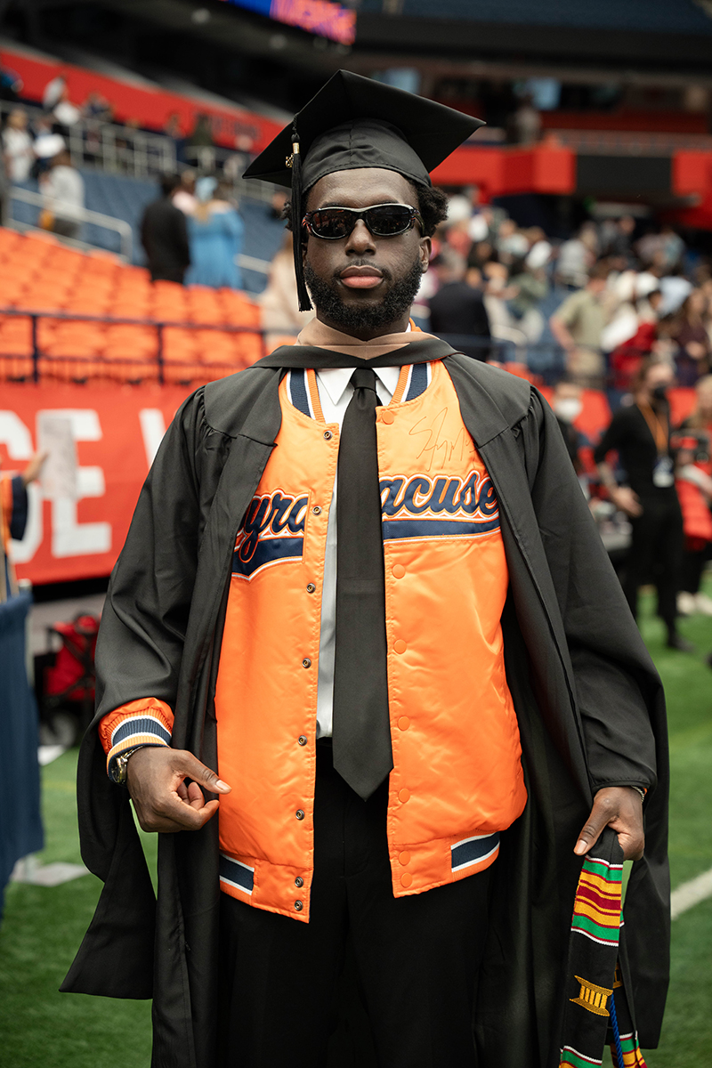 A person wearing a graduation cap and gown over an orange jacket with the word 'Syracuse' written on it. The person is standing on a field in the JMA Wireless Dome, with blurred spectators in the background.