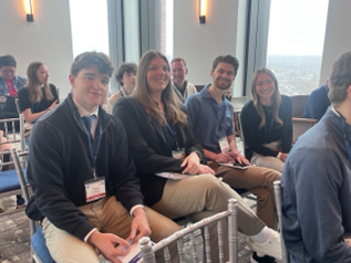 A group of people seated in chairs in a conference room with large windows. They are dressed in business casual attire and wearing name tags, suggesting they are attending an event or meeting. A cityscape is visible through the windows in the background.