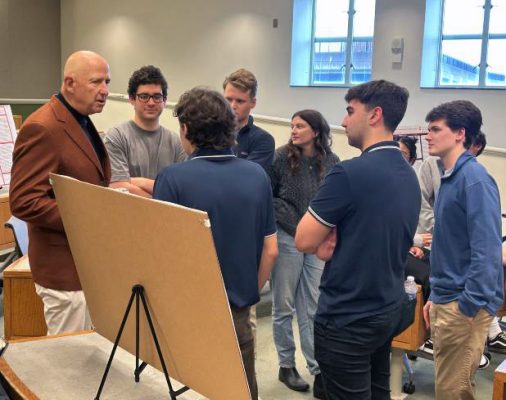 A group of seven people stand in a semi-circle around a presentation board on an easel, engaged in a discussion or presentation. The setting is an indoor room with natural light streaming through windows in the background.