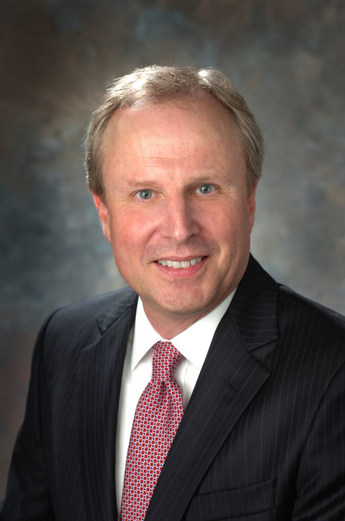 A person is wearing a dark pinstripe suit, a white dress shirt, and a red patterned tie. The background is blurred with shades of blue and gray.