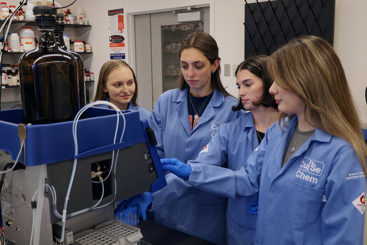 The image shows four individuals wearing blue lab coats with "Cuse Chem" logos gathered around a piece of laboratory equipment. The equipment has a large brown bottle on top and several tubes connected to it. The background includes shelves with various containers and a cabinet with glass doors.