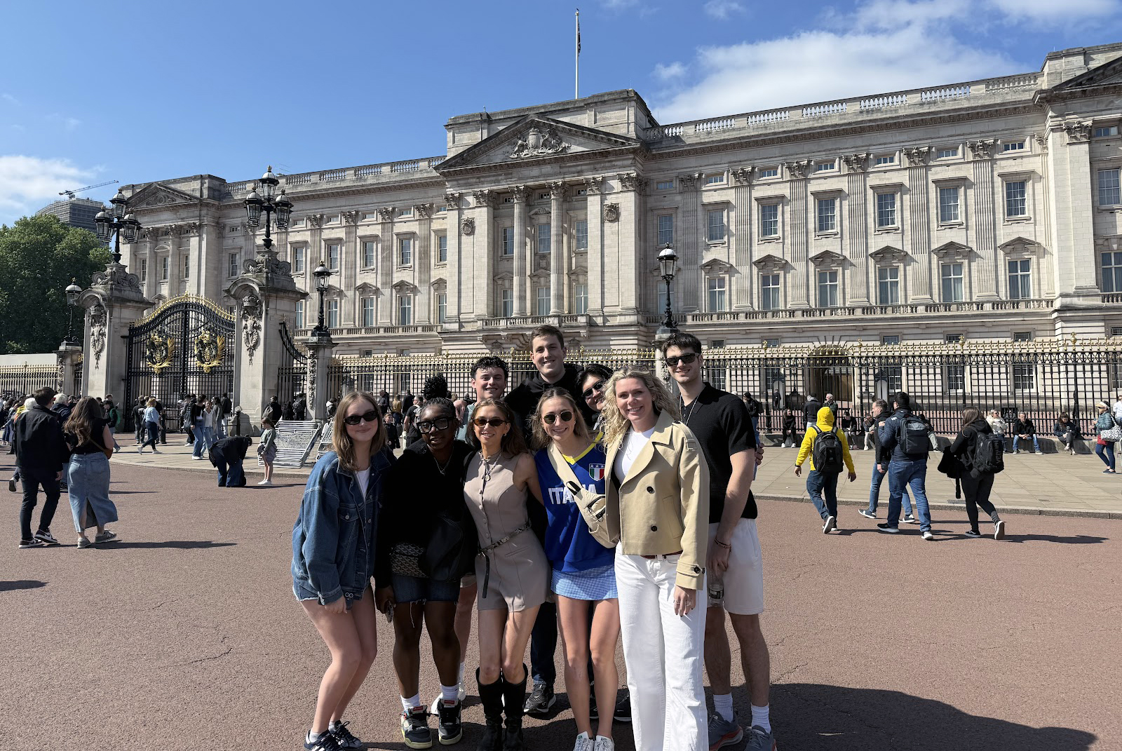 Students gather in front of Buckingham Palace in London, with the grand facade of the palace featuring numerous windows and ornate architectural details under a partly cloudy sky.