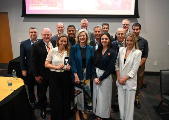 Group of twelve people posing indoors in front of a projection screen displaying a presentation, with some individuals holding items like a smartphone, suggesting an event or conference setting.