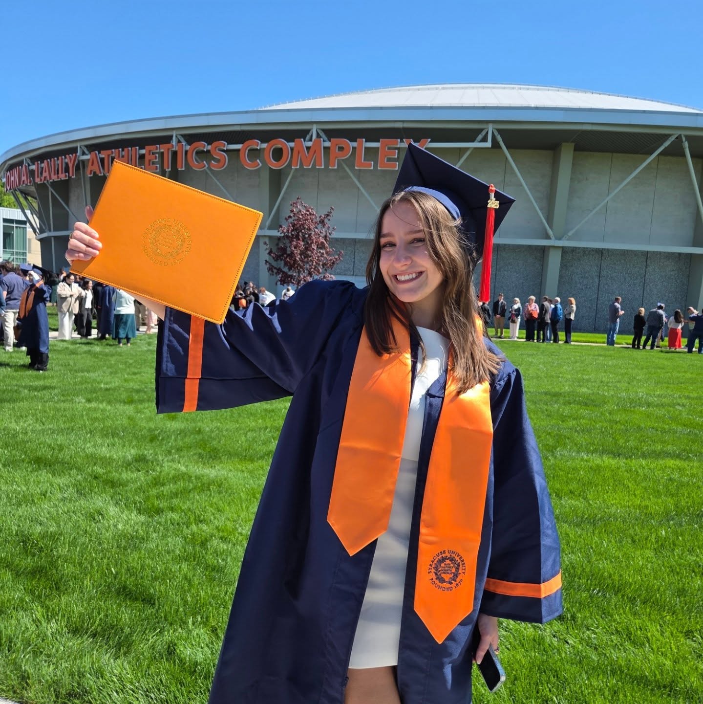 A person in graduation attire holds up a diploma in front of the John A. Lally Athletics Complex. The background features a rounded building, other people and trees under a clear blue sky.