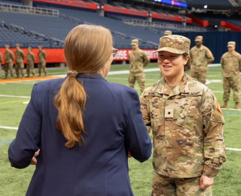 A person in a blue blazer shakes hands with Army ROTC cadet Hannah Kang in military uniform. In the background, more cadets stand in formation on an indoor sports field. The cadet has the name tag "KANG" and "U.S. ARMY" on the uniform.