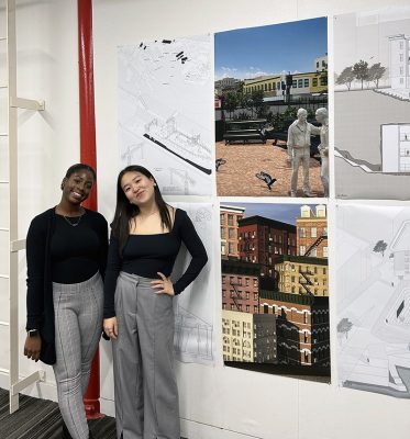 Two individuals stand in front of a wall displaying six architectural drawings and renderings. The drawings include detailed plans and perspectives of buildings and urban spaces. The two individuals are dressed in dark tops and light-colored pants.