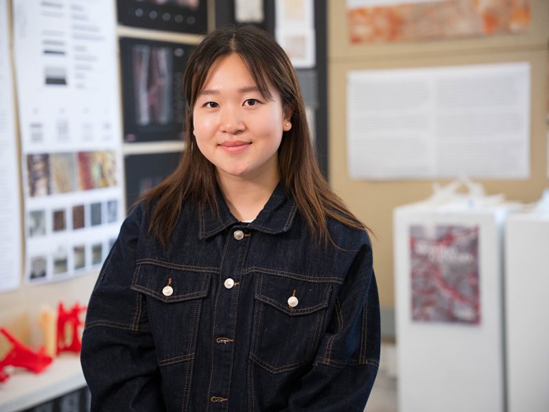 A student stands in front of a display of posters and images. She is wearing a dark denim jacket with white buttons. The background features various posters for an architecture class studio, some with text and others with images, displayed on walls and boards.
