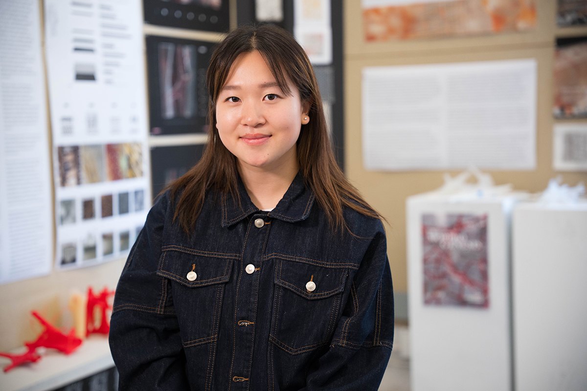A student stands in front of a display of posters and images. She is wearing a dark denim jacket with white buttons. The background features various posters for an architecture class studio, some with text and others with images, displayed on walls and boards.