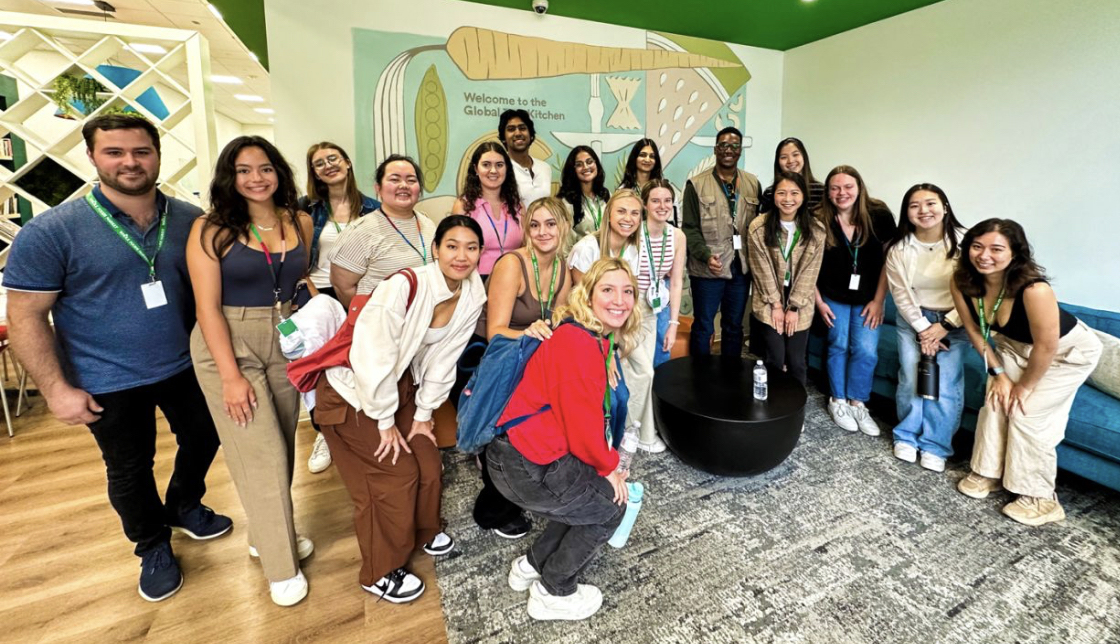 A group of interns are standing and sitting in a room with green walls and wooden flooring. They are posing for a photo in front of a wall mural that features vegetables and the text "Welcome to the Global Kitchen" at Whole Foods Market Headquarters. There is a black round table in the center with a water bottle on it, and some people are holding papers or bags.