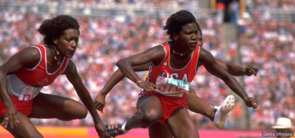 three runners have intense looks on their faces as they compete in a track-and-field event in the Olympics. In the background, spectators can be viewed in the stands.
