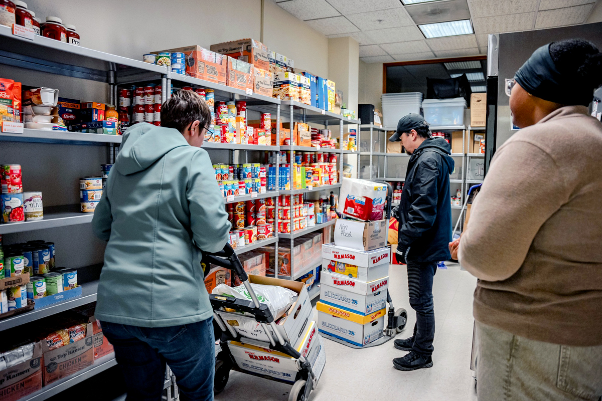Three individuals in a storage room or pantry filled with shelves stocked with canned goods, cereal boxes, and other non-perishable food items. One person pushes a cart loaded with 'Kleenex' boxes, another stands beside a stack of similar boxes, and a third person observes the activity.