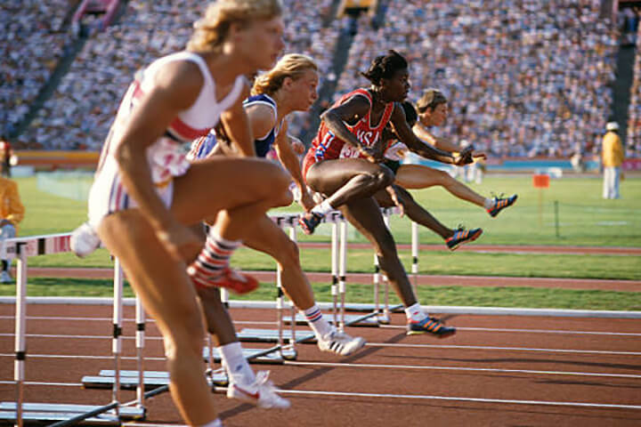 five athletes are pictured jumping over hurdles at an Olympic event. They are competing on a track, and there is green grass and stands full of spectators behind them.