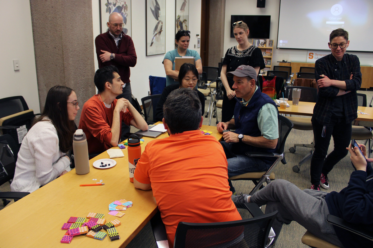 A group of eight people are gathered around a table in a meeting or collaborative work session. Some are seated while others are standing. The table has various items on it, including colorful hexagonal tiles, notebooks, cups, and a water bottle. In the background, there is a projector screen displaying an image or presentation. The room has beige walls and framed pictures hanging on them.