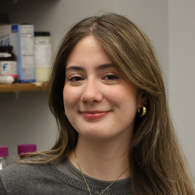The image shows a person with long brown hair wearing a gray sweater and gold hoop earrings. The background includes shelves with various containers, including a white bottle with a blue label and some purple-capped bottles.